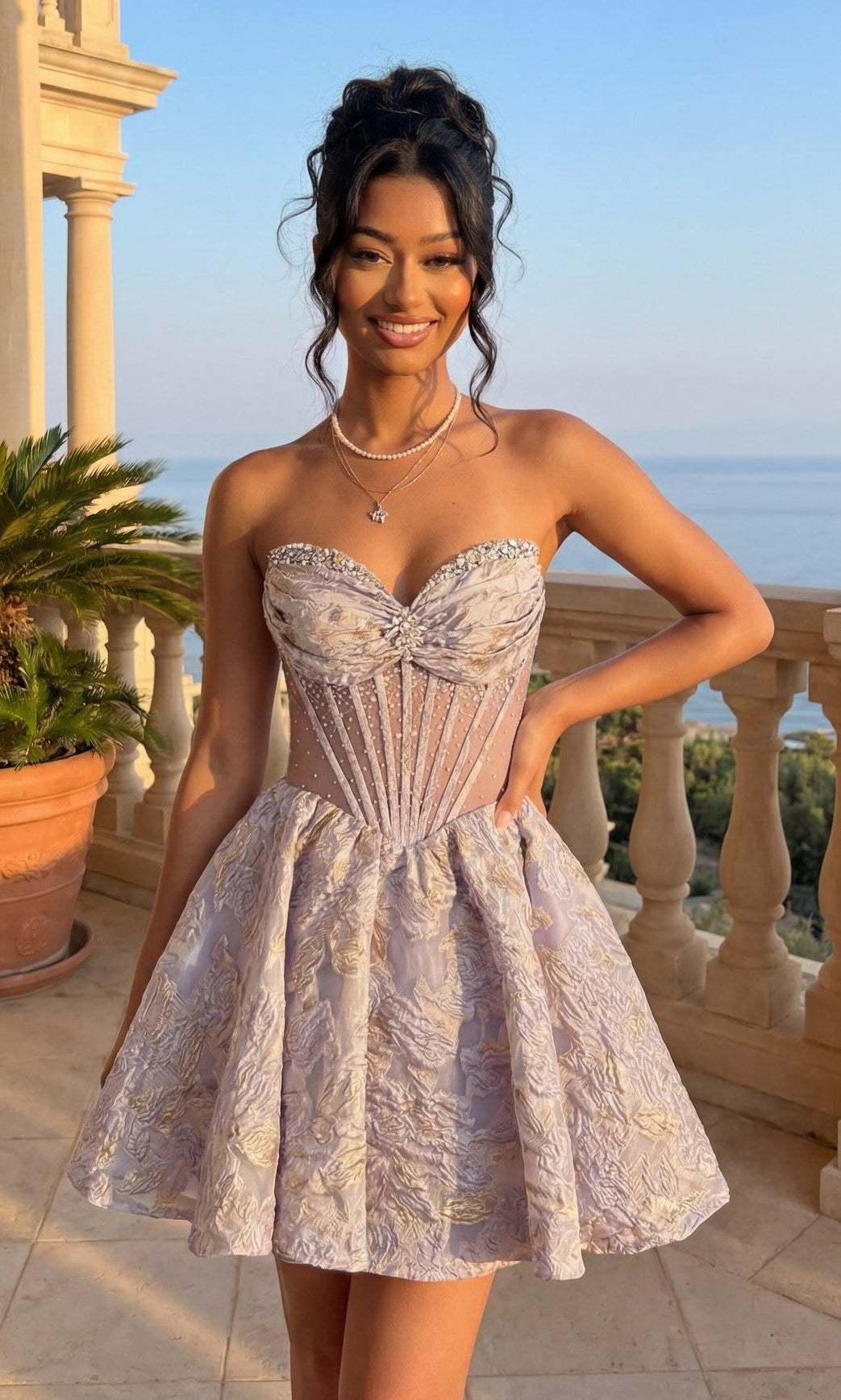 A woman in a lilac strapless Short Homecoming Dress (Amelia Couture 8075S) with a corset bodice stands on a stone balcony with plants and columns, gazing at the ocean.